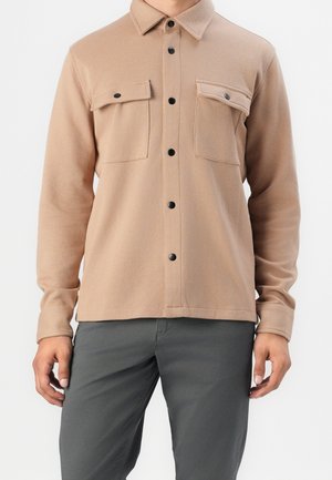 Chemise beige à boutons, style veste, avec deux poches poitrine à rabat, portée avec un pantalon gris foncé, vue du cou jusqu'aux hanches.