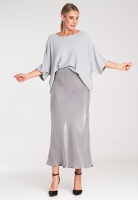 Hellgraues Strickoberteil mit weiten Ärmeln, über einem grauen Satin-Rock mit Unterrock geschichtet. Schwarze Riemchen-Absätze vervollständigen das Outfit. Einfaches Design und weiche Texturen.