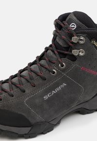 Bota de senderismo de ante negro con cordones rojos y negros, ojales de metal y suela de goma texturizada. Logo "SCARPA" destacado.