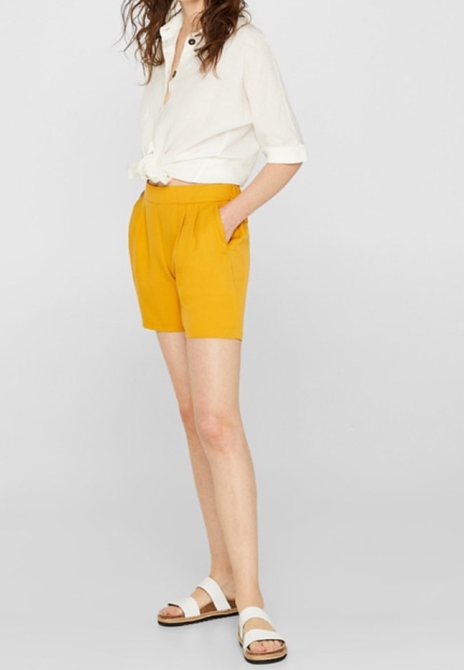 Femme debout les mains dans les poches, portant un short plissé jaune moutarde, une chemise blanche aux manches retroussées nouée à la taille et des sandales blanches.