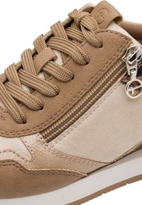 Gros plan d'une basket beige et marron avec des lacets tissés, une fermeture éclair latérale avec tirette en métal, et des détails cousus sur des matériaux doux en daim et cuir.
