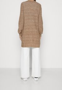 Brązowy sweter z dzianiny z długimi, bufiastymi rękawami, o prążkowanej fakturze. Do tego białe spodnie o szerokich nogawkach i białe trampki.