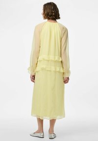 Largo vestido amarillo hecho de tela transparente con acentos con volantes, mangas largas y un diseño fluido, combinado con zapatos planos plateados.