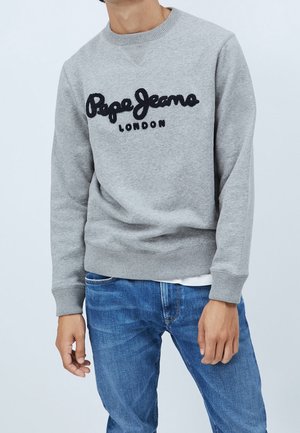 Mężczyzna w szarej bluzie z napisem "Pepe Jeans London" i niebieskich jeansach na tle jasnej, jednolitej ściany.