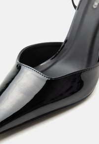 Μαύρο παπούτσι γόβας από λουστρίνι με μυτερή μύτη και λουρί στο αστράγαλο, σε κοντινή λήψη σε λευκό φόντο.