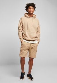 Hoodie beige con cordini, grande tasca frontale e pantaloni cargo abbinati con tasche laterali. Indossato con sneakers nere e arancioni.