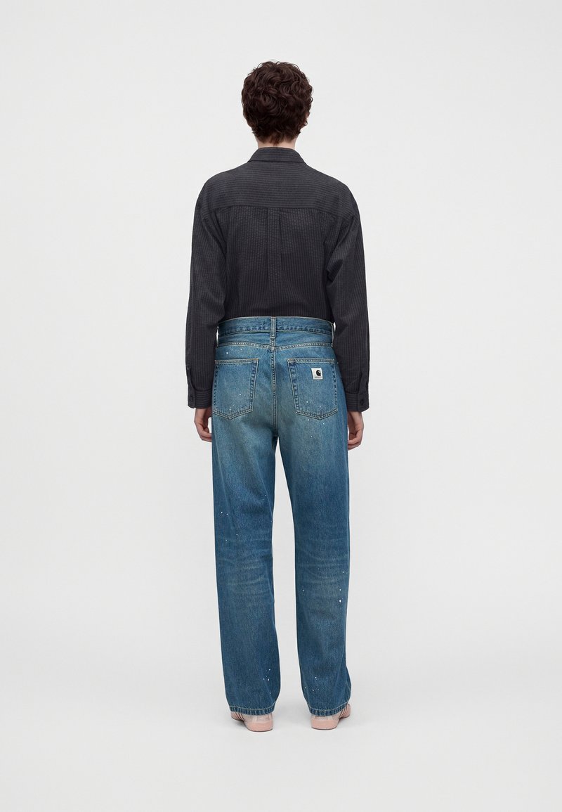 Persona con capelli corti e ricci che indossa una maglia nera a maniche lunghe e jeans blu larghi con tasche posteriori e patch con logo, in piedi su uno sfondo semplice.