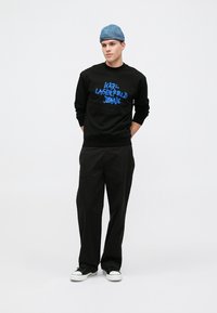 Čierny sveter s okrúhlym výstrihom a modrou grafikou "KARL LAGERFELD JEANS", skombinovaný s voľnými čiernymi nohavicami a bielymi teniskami, doplnený svetlomodrou čiapkou.