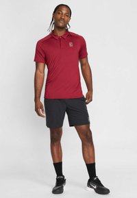 Polo vermelha de manga curta com colarinho, combinada com calções pretos. Apresenta um pequeno logótipo no peito e sapatos desportivos pretos.