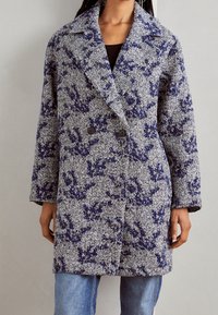 Vrouw in een lange, getextureerde wollen jas met een blauw-wit bloemenpatroon over een zwarte top en lichtblauwe jeans.