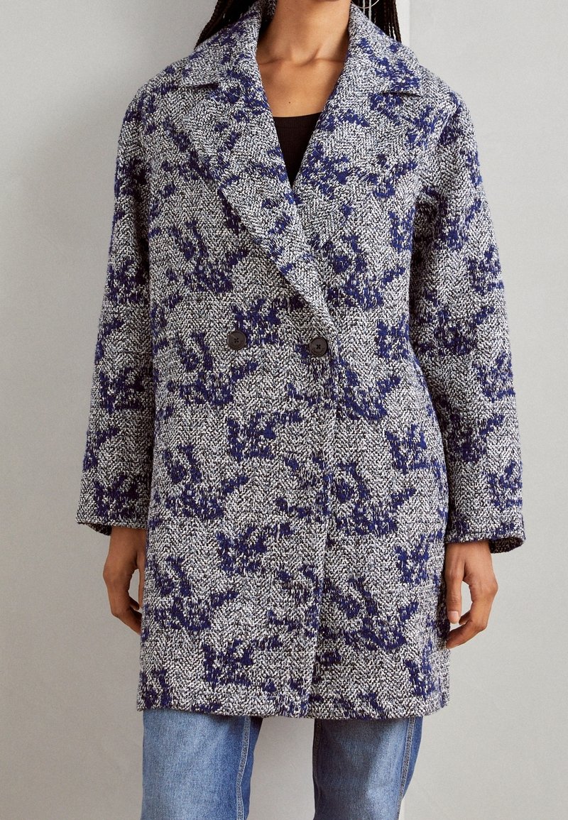 Vrouw in een lange, getextureerde wollen jas met een blauw-wit bloemenpatroon over een zwarte top en lichtblauwe jeans.