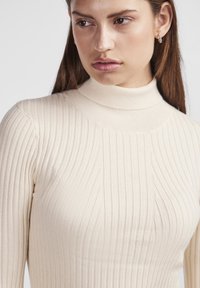 Beige ribbad polokrage med en åtsittande design, med vertikala ränder och hög halsringning. Tyget ser mjukt och strukturerat ut.