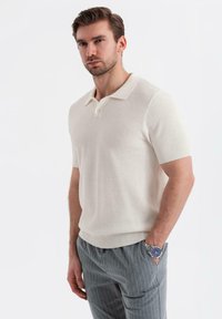 Maglione polo a maniche corte color avorio, caratterizzato da un motivo a maglia testurizzata, con polsini a costine e colletto piatto. Indossato con pantaloni a righe grigie.