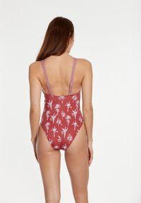 Eénstuk zwemkleding in een rode tint met een witte palmboomprint. Heeft brede patroonstraps en een lage rugontwerp.