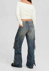 Wyblakłe niebieskie dżinsy cargo z dużymi bocznymi kieszeniami, szerokimi nogawkami i postrzępionymi zakończeniami, w połączeniu z białym, krótkim swetrem.