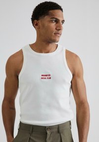 Hvid ærmeløs tanktop med ribbet tekstur, der har rød broderet tekst "PROHIBITED SOCIAL CLUB" på brystet, parret med khakifarvede bukser.