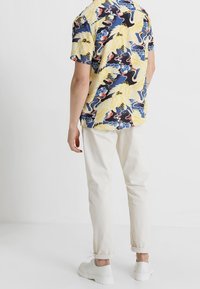 Chemise à manches courtes jaune avec un motif de grue bleu, rouge et blanc. Associée à un pantalon de couleur claire et des baskets blanches.