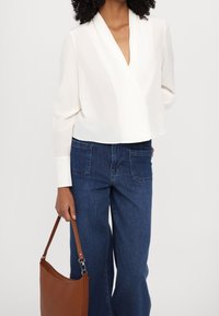 Blusa blanca de estilo envolvente con mangas largas, combinada con unos jeans de pierna ancha azules. La persona lleva un bolso de hombro de cuero marrón.