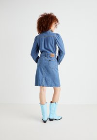Vestido de ganga com mangas bufantes compridas, cintura com cinto e fenda nas costas. Usado com botas de cowboy azuis. Logótipo da Lee na parte de trás.
