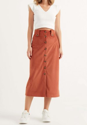 Vrouw draagt een midi-rok van ribfluweel in roestkleur met knopen aan de voorkant, een witte mouwloze top met V-hals en witte sneakers, staand tegen een effen achtergrond.