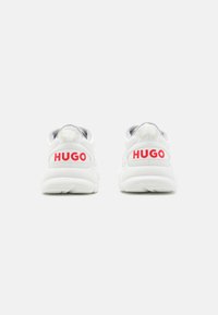 White athletic shoes with a textured surface, featuring red "HUGO" branding on the heel. Rounded shape with a padded tongue and collar.