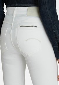 Vaqueros de mezclilla blanca con tela texturizada, cintura alta y costuras sutiles en los bolsillos traseros. Cuenta con un bolsillo con cremallera para almacenamiento.