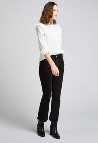 Blouse blanche en broderie anglaise avec des accents volantés et des manches retroussées, associée à un pantalon en velours côtelé noir et ajusté, ainsi qu'à des bottines noires à talons.