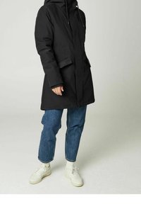 Person wearing black hooded coat, blue jeans, white socks, and white sneakers, standing against a plain light background.