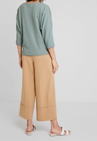 Kvinna står med ryggen vänd, klädd i en ljusblå, texturerad långärmad tröja, vida beige cropped-byxor och vita sandaler.