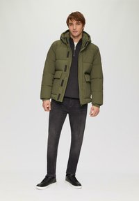 Olivgrüne Steppjacke mit Kapuze, Reißverschluss vorn und zwei Vordertaschen. Getragen über einem dunkelgrauen Oberteil mit schmalen, dunklen Jeans und schwarzen Sneakers.