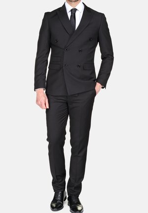 Homme portant un costume noir croisé avec une chemise blanche, une cravate noire, un pantalon noir assorti et des chaussures de ville noires, debout avec une main dans la poche.