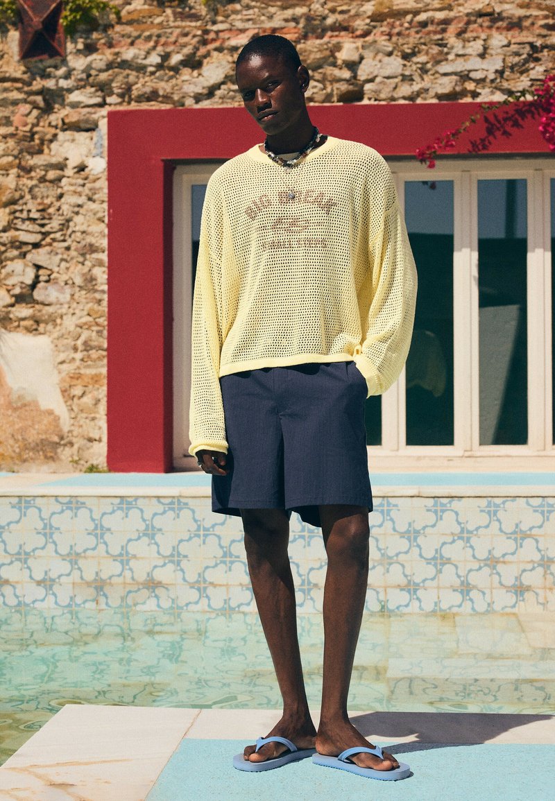 Personne portant un pull en maille jaune, un short foncé et des tongs bleues, debout à côté d'une piscine carrelée avec un mur en pierre et une fenêtre en arrière-plan.