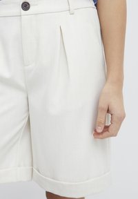 Pantaloni beige plissettati realizzati in tessuto leggero, con dettaglio a bottone e tasche laterali con orlo arrotolato alle aperture delle gambe.