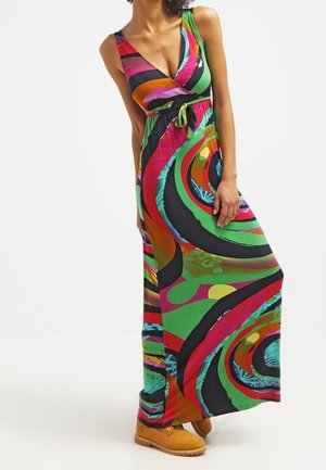 Femme portant une robe maxi sans manches colorée avec des motifs circulaires abstraits et des bottes beige à lacets, posant devant un fond blanc.