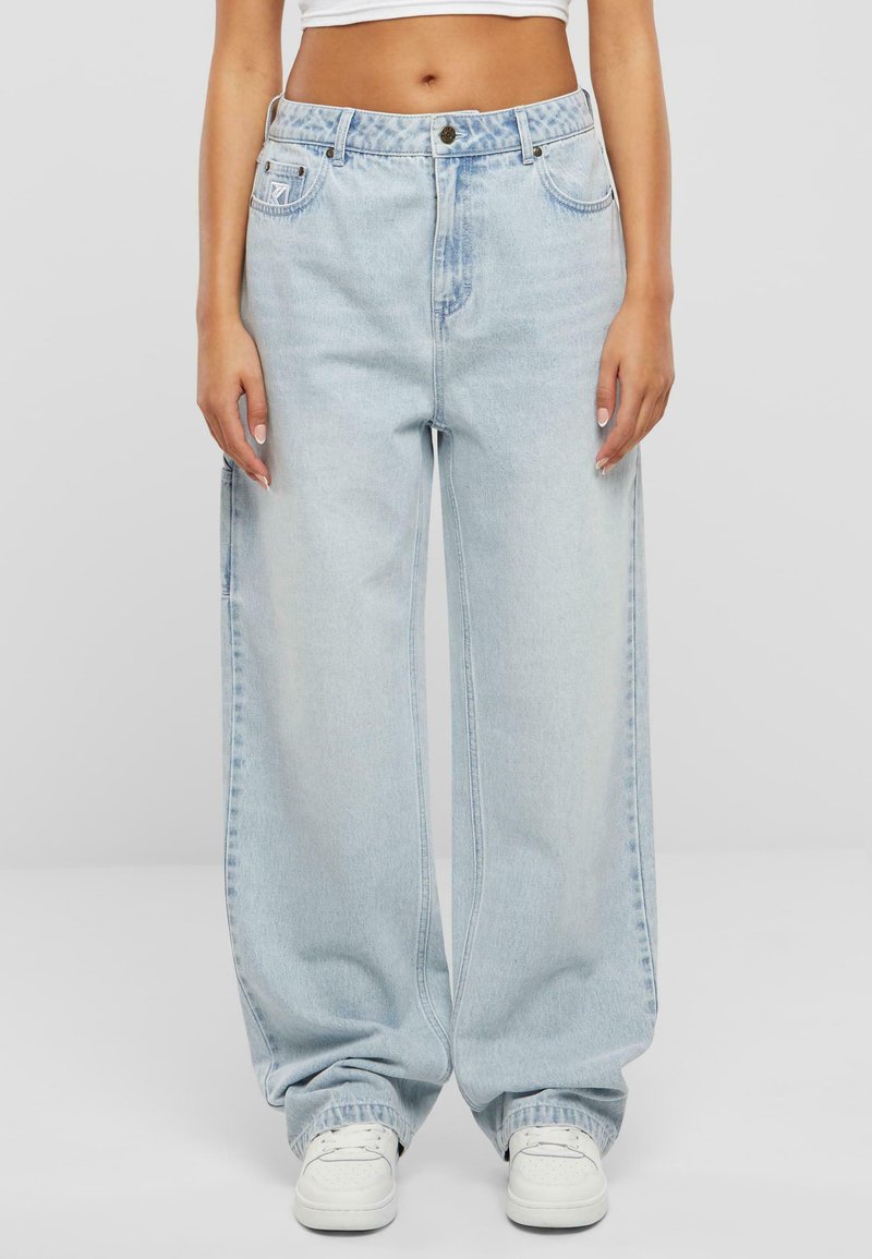 Jean large bleu clair en denim, avec une taille haute, des poches avant et une fermeture à boutons, associés à des baskets blanches.