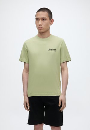 Jeune homme portant un t-shirt Barbour vert clair et un short noir, debout devant un fond blanc uni, regardant sur le côté.