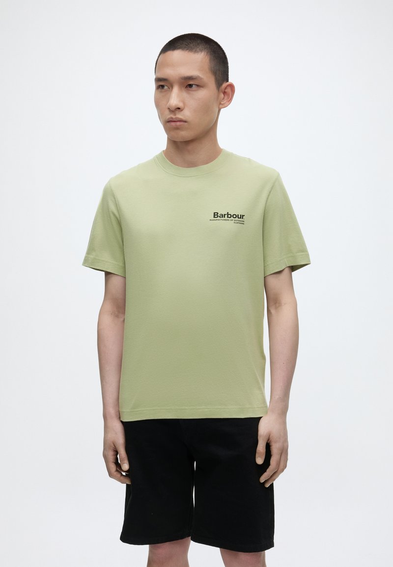 Νέος άνδρας που φοράει ένα ανοιχτό πράσινο t-shirt Barbour και μαύρα σορτς, στέκεται μπροστά σε λευκό φόντο και κοιτάζει στο πλάι.