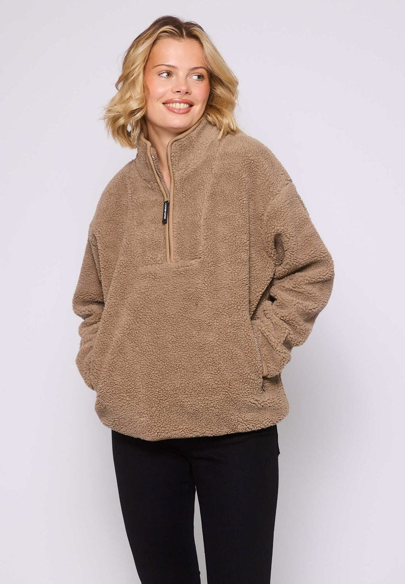 Uśmiechnięta kobieta z krótkimi blond włosami, ubrana w beżowy polar z zamkiem błyskawicznym w kołnierzu i czarne spodnie, z rękami w kieszeniach, patrząca w prawo.