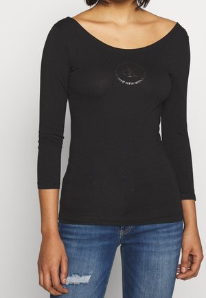 Kobieta ubrana w czarną, dopasowaną bluzkę Calvin Klein z długim rękawem i szerokim dekoltem oraz niebieskie, podarte dżinsy, stojąca na jednolitym tle.