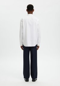 Άσπρο πουκάμισο με κουμπιά και μακριά μανίκια, με κλασικό γιακά και στρογγυλεμένη τελείωμα, συνδυασμένο με σκούρο μπλε παντελόνι με φαρδιά πόδια και μαύρα παπούτσια.