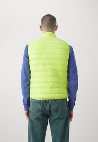 Prsluk od limunasto zelene boje s kvačkastim dizajnom, s podignutim ovratnikom i bez rukava, u kombinaciji s plavom košuljom dugih rukava i trapericama.