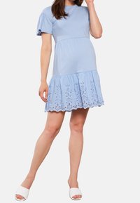 Vaaleansininen puuvillamekko, jossa on lyhyet hiha, kerrostettu muotoilu ja silmänrei'ityksellä koristeltu helma. Yhdistetty valkoisiin tilkkulampaankengiin.