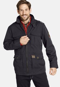 Navy utility jacket with zip front, multiple pockets, and shoulder epaulets. Features a red inner lining and a woven brand label at the hem.