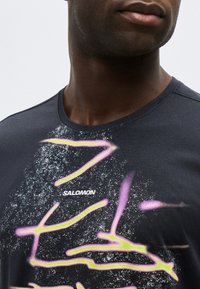 Κοντινό πλάνο ενός ατόμου που φοράει μαύρο μπλουζάκι Salomon με ροζ και κίτρινες αφηρημένες γραμμές και λευκό βουρτσισμένο σχέδιο στο στήθος.