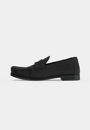 Čierny kožený slip-on mokasína s nízkym podpätkom, šitím moc toe a širokým remienkom cez hornú časť na hladkom svetlom pozadí.