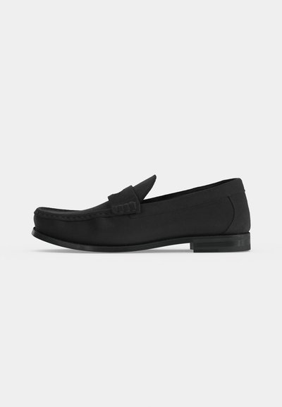 Μαύρο δερμάτινο μοκασίνι slip-on με χαμηλό τακούνι, ραφή τύπου moc toe και φαρδιά λωρίδα στο πάνω μέρος σε απλό ανοιχτό φόντο.