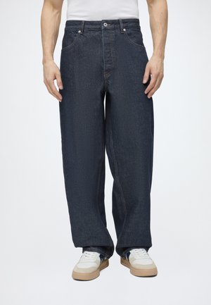 Mand iført mørkeblå denimjeans med brede ben, forlommer og hvide sneakers på ensfarvet baggrund.