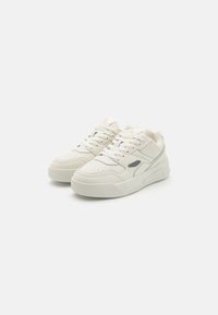 White athletic sneakers with a textured leather upper, perforated toe box, and contrasting dark detailing on the side. Cushioned sole and lacing.