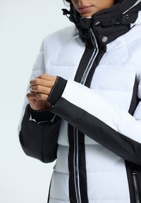 Gevoerde jas met een zwart-wit kleurenschema, textuurstof, verstelbare capuchon en duimopening in de mouwen. Rits sluiting aan de voorkant.