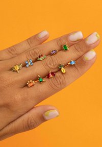 Dieci anelli di vari colori e forme esposti su una mano: accenti dorati, gemme in giallo, blu, verde, rosso e viola su uno sfondo arancione.